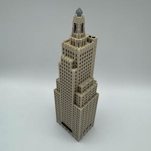 May include: A beige, miniature skyscraper model with a detailed design. The building has multiple tiers and a decorative top. The model is made of a light-colored material and features numerous small windows. The overall design is reminiscent of classic architecture.