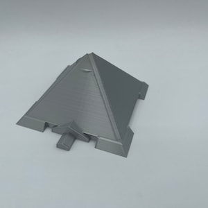 Puede incluir: Un objeto gris en forma de pir&aacute;mide con una base plana y lados angulados. La pir&aacute;mide tiene una superficie texturizada y peque&ntilde;as extensiones rectangulares en la base. El objeto est&aacute; sobre un fondo blanco.