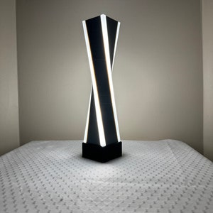 May include: A black and white modern table lamp with a twisting design. The lamp has a white light source and is on a white table.