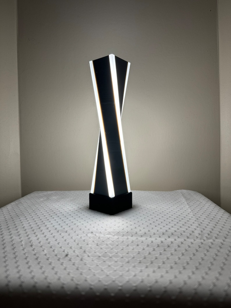 May include: A black and white modern table lamp with a twisting design. The lamp has a white light source and is on a white table.