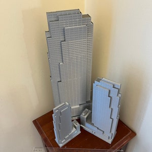 May include: Three gray 3D printed models of skyscrapers, the tallest is about 12 inches tall, the middle is about 8 inches tall, and the smallest is about 6 inches tall. The models are sitting on a wooden table with a ruler.