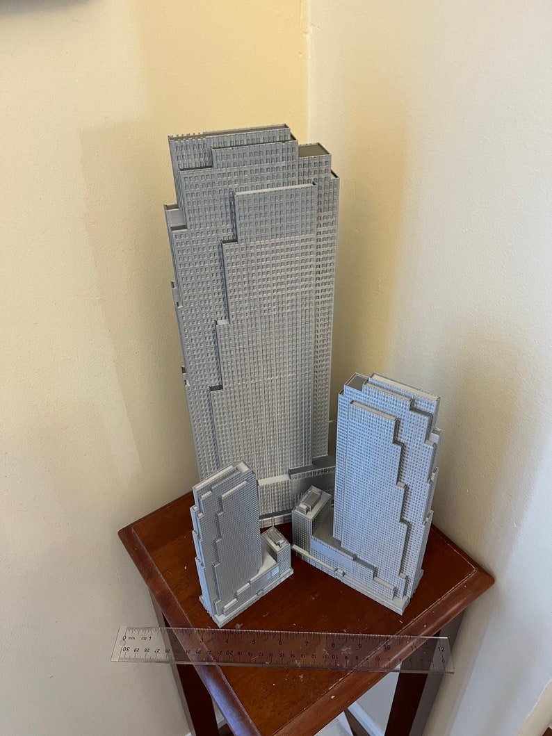 May include: Three gray 3D printed models of skyscrapers, the tallest is about 12 inches tall, the middle is about 8 inches tall, and the smallest is about 6 inches tall. The models are sitting on a wooden table with a ruler.