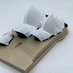 May include: A white 3D printed model of the Sydney Opera House. The model is on a tan base.