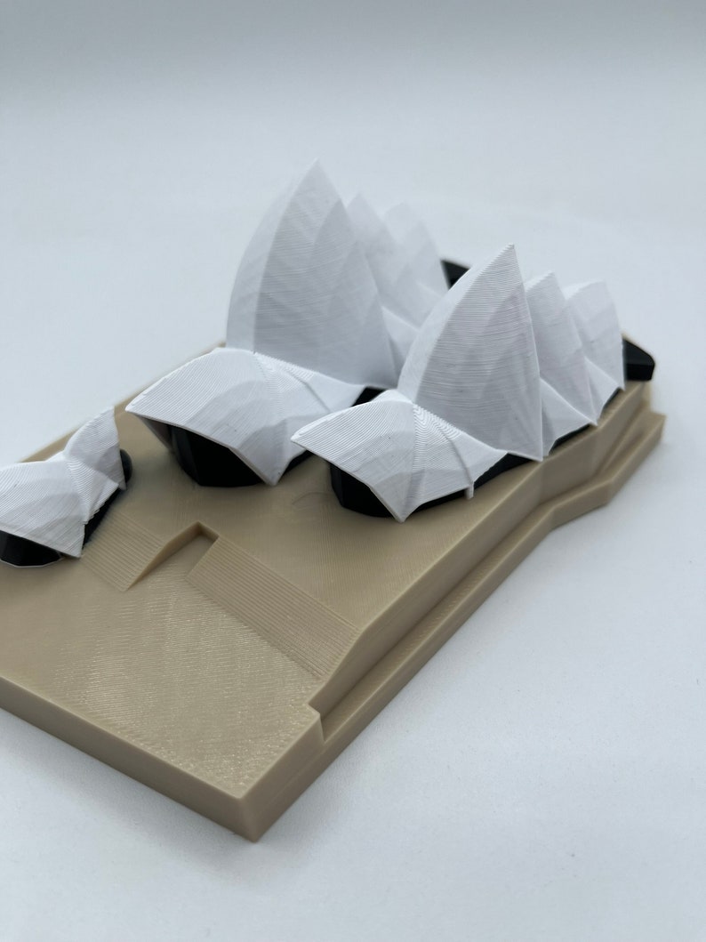 May include: A white 3D printed model of the Sydney Opera House. The model is on a tan base.
