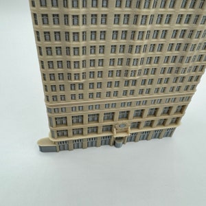 May include: A beige miniature building model with a detailed facade featuring many windows. The model has a slightly curved shape and a flat roof.