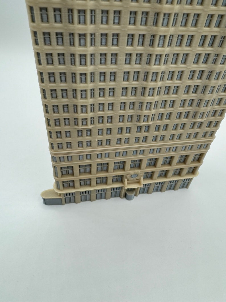 May include: A beige miniature building model with a detailed facade featuring many windows. The model has a slightly curved shape and a flat roof.