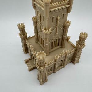 May include: A beige 3D-printed model of a castle with multiple turrets and architectural details. The model features a central tower and several smaller towers, showcasing intricate design elements and a detailed facade. The overall structure is a miniature representation of a historical building.