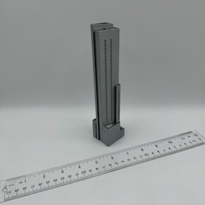 May include: A gray, 3D-printed model of a skyscraper, featuring numerous windows and a rectangular base. A clear ruler with inch markings is placed next to the building. The model appears to be approximately 4 inches tall.