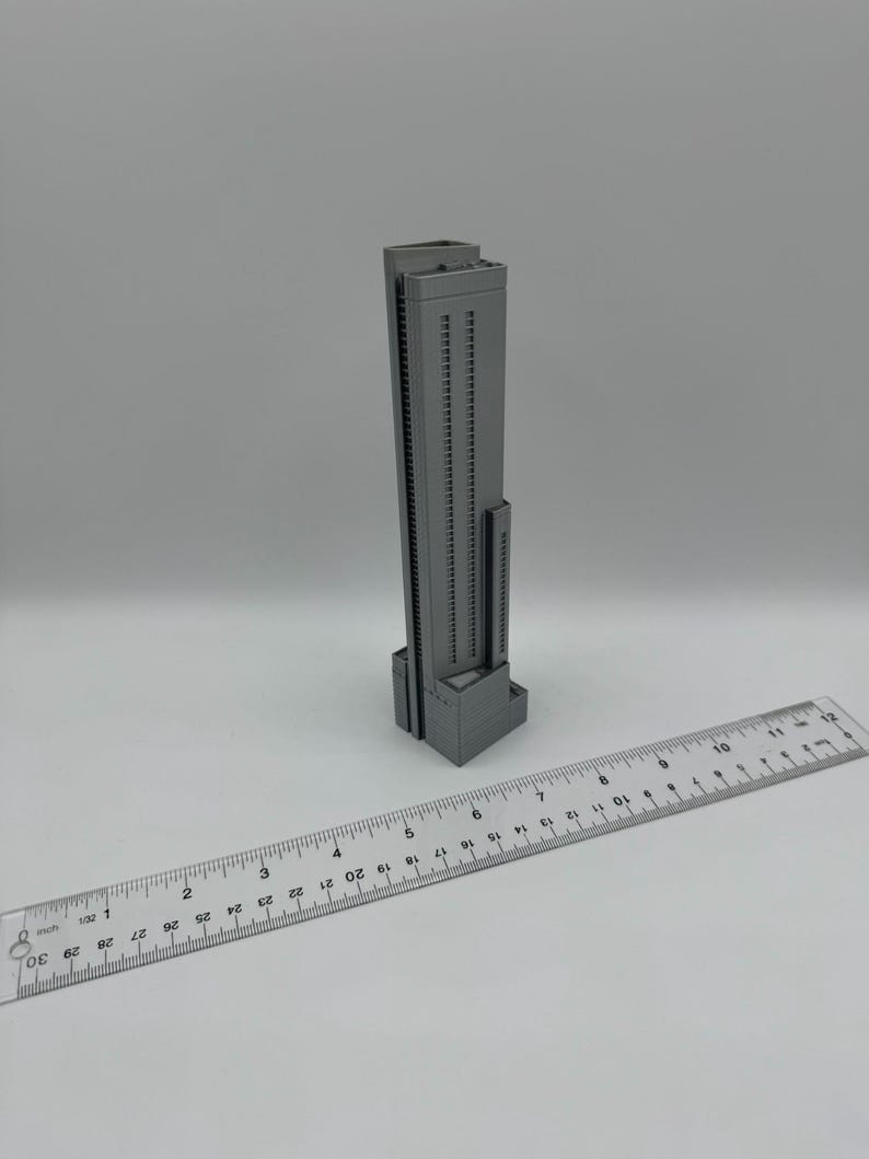 May include: A gray, 3D-printed model of a skyscraper, featuring numerous windows and a rectangular base. A clear ruler with inch markings is placed next to the building. The model appears to be approximately 4 inches tall.