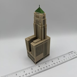 May include: A beige architectural model of a skyscraper with a green roof. The model has detailed windows and a rectangular base. A ruler is placed next to the model, showing measurements in inches.