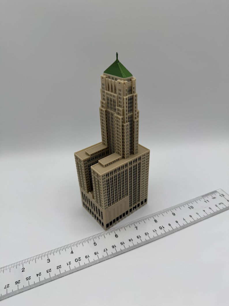 May include: A beige architectural model of a skyscraper with a green roof. The model has detailed windows and a rectangular base. A ruler is placed next to the model, showing measurements in inches.