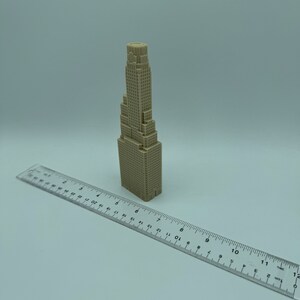 May include: A beige 3D printed model of a skyscraper building. The model is about 6 inches tall and is sitting on a white surface next to a ruler.