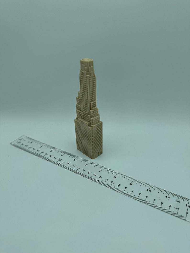 May include: A beige 3D printed model of a skyscraper building. The model is about 6 inches tall and is sitting on a white surface next to a ruler.