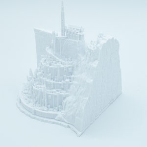 May include: A white 3D printed model of a fantasy castle built on a cliffside. The castle has a tall tower and many smaller buildings.