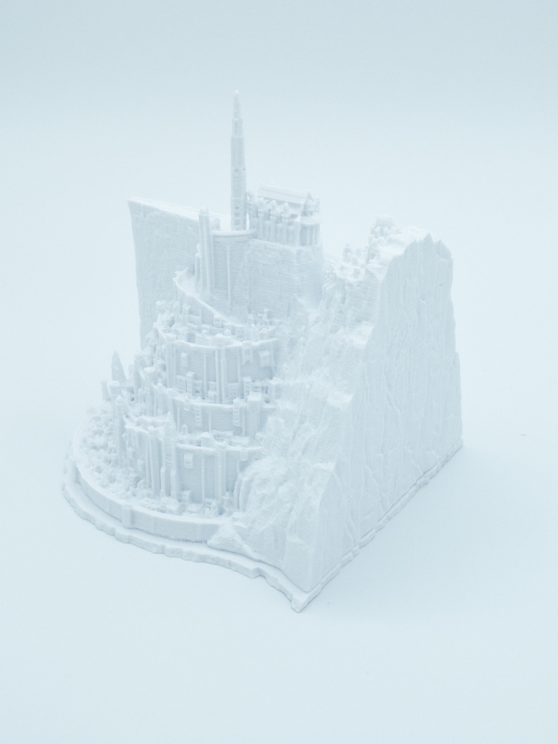 May include: A white 3D printed model of a fantasy castle built on a cliffside. The castle has a tall tower and many smaller buildings.