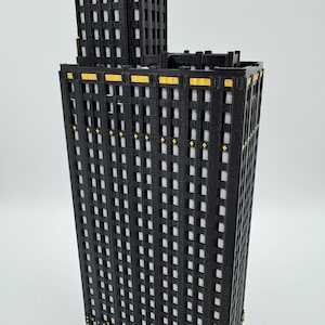 Op de afbeelding: Een 3D-geprint model van een zwarte en gouden wolkenkrabber. Het gebouw heeft veel ramen en gouden accenten. Het model staat tegen een witte achtergrond.