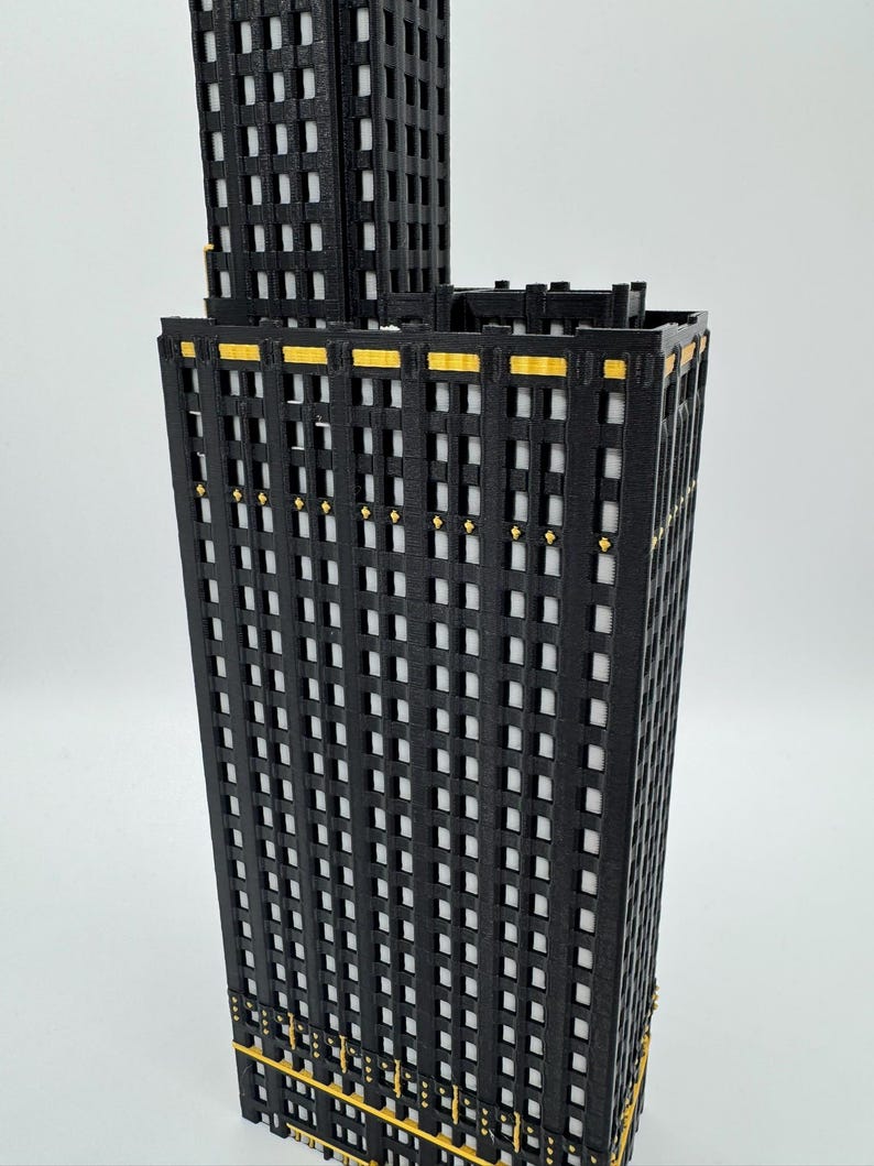 Op de afbeelding: Een 3D-geprint model van een zwarte en gouden wolkenkrabber. Het gebouw heeft veel ramen en gouden accenten. Het model staat tegen een witte achtergrond.