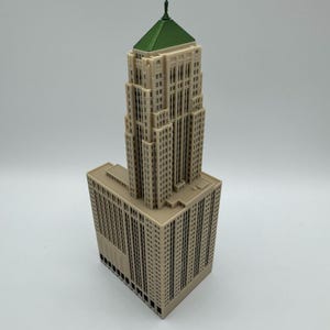 May include: A beige architectural model of a skyscraper with a green roof and a small statue on top. The building has many windows and is a detailed replica of a famous structure.