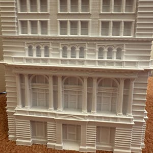 Op de afbeelding: Een wit 3D-geprint model van een gebouw met meerdere ramen en decoratieve details. Het gebouw heeft een plat dak en een eenvoudige gevel.