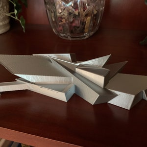 May include: A silver-colored, 3D printed model of a modern building with a geometric design. The model has a series of angled, flat surfaces that create a unique and abstract shape.