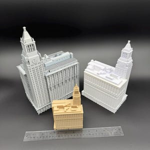 May include: Three miniature building models in shades of gray and tan are displayed against a dark background. The models include detailed architectural features, with one featuring a clock tower. A ruler is placed in front of the buildings.