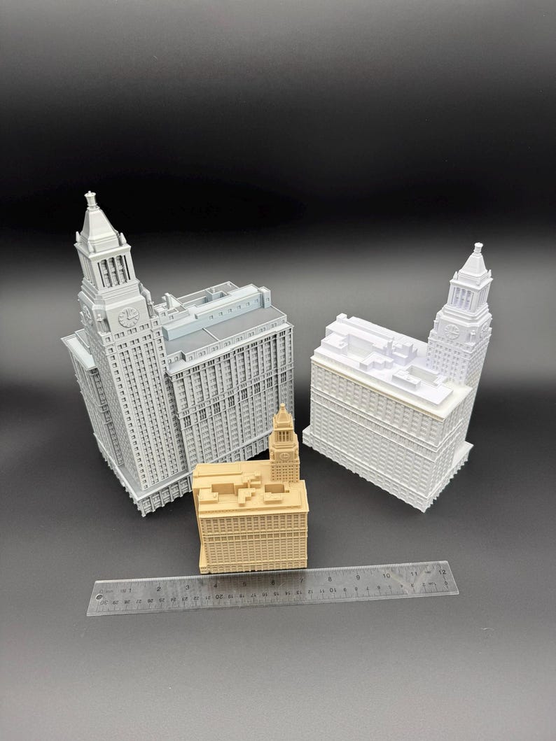 May include: Three miniature building models in shades of gray and tan are displayed against a dark background. The models include detailed architectural features, with one featuring a clock tower. A ruler is placed in front of the buildings.
