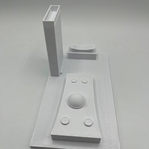 K&ouml;nnte beinhalten: Wei&szlig;es 3D-gedrucktes Modell einer Stadtlandschaft mit einem hohen Geb&auml;ude, einem kuppelf&ouml;rmigen Bauwerk und einem gebogenen Geb&auml;ude. Das Modell steht auf einem wei&szlig;en Sockel.
