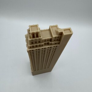 May include: A beige architectural model of a skyscraper. The building has a rectangular shape with many windows and detailed rooftop features. The model is made of a solid material and sits on a white surface.