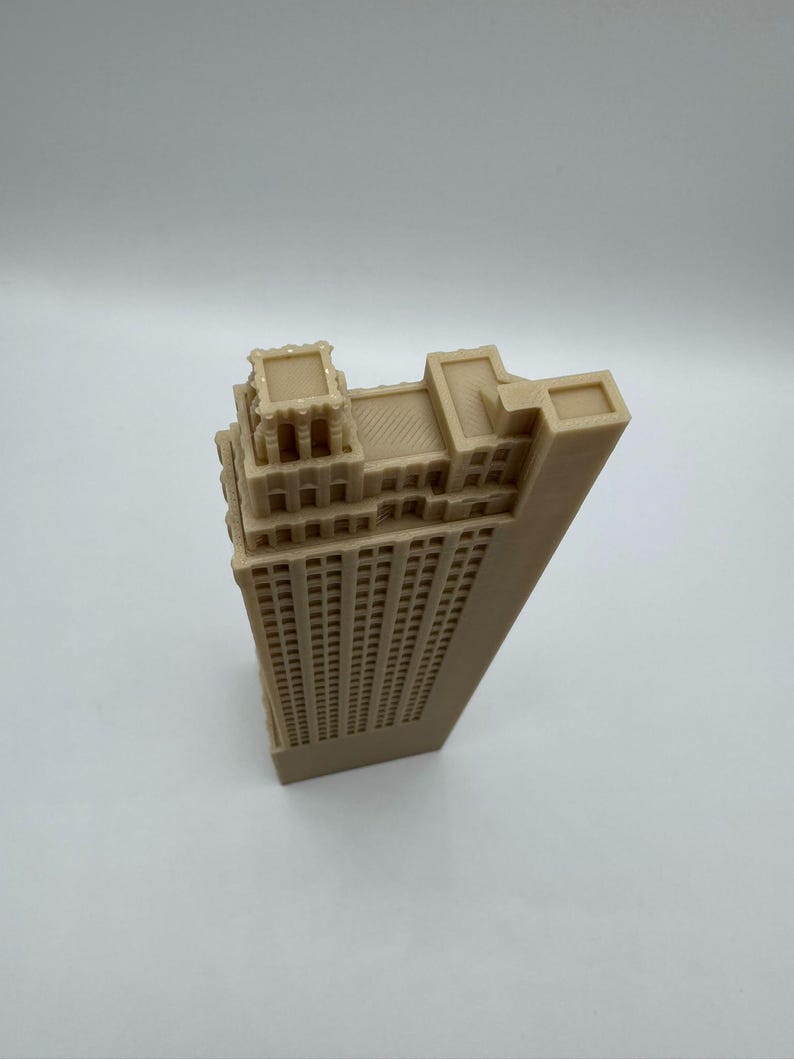 May include: A beige architectural model of a skyscraper. The building has a rectangular shape with many windows and detailed rooftop features. The model is made of a solid material and sits on a white surface.