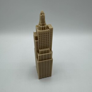 May include: A tan-colored miniature skyscraper model with intricate architectural details. The model features multiple levels, windows, and a detailed top section with a dome. The building is a representation of a classic architectural style.