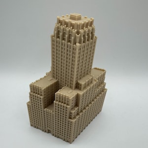 May include: A tan-colored architectural model of a skyscraper. The model has a tiered design with a central tower and multiple lower sections. The building features detailed window patterns and a flat roof.