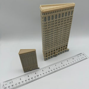 May include: Two miniature models of skyscrapers, one taller than the other, both made of beige plastic with detailed window and door features. The models are placed on a white surface with a ruler for scale.