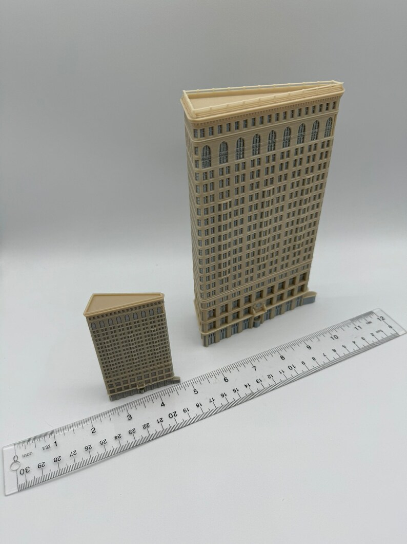 May include: Two miniature models of skyscrapers, one taller than the other, both made of beige plastic with detailed window and door features. The models are placed on a white surface with a ruler for scale.