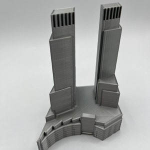 Op de afbeelding: Een 3D-geprint model van een moderne wolkenkrabber met twee torens die met elkaar verbonden zijn door een lagere structuur. Het model is gemaakt van grijs plastic en heeft een gedetailleerd ontwerp.