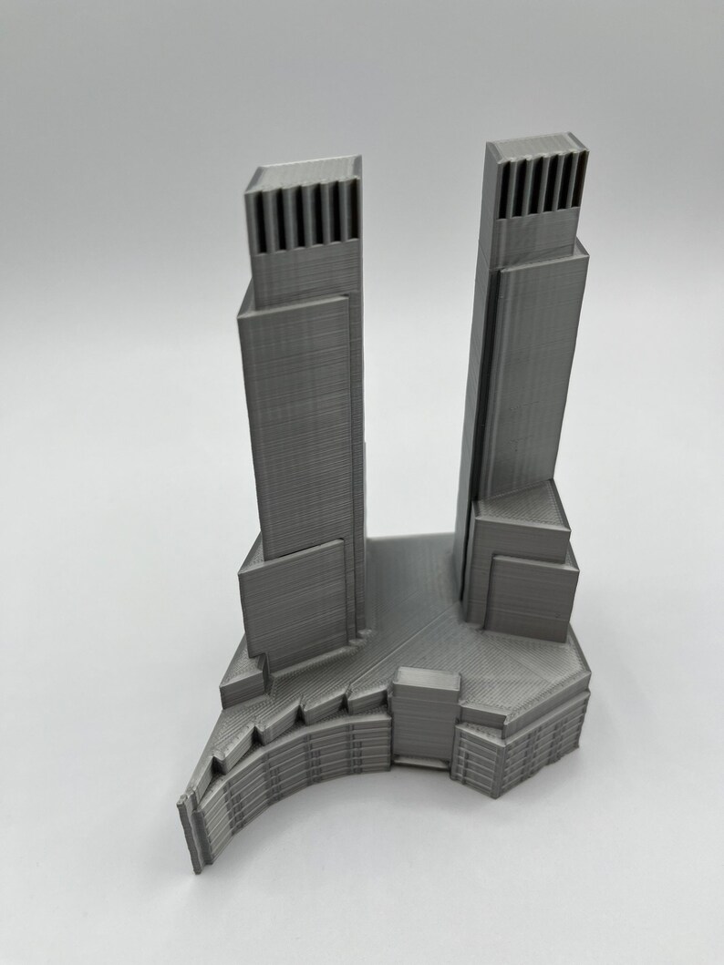 Op de afbeelding: Een 3D-geprint model van een moderne wolkenkrabber met twee torens die met elkaar verbonden zijn door een lagere structuur. Het model is gemaakt van grijs plastic en heeft een gedetailleerd ontwerp.