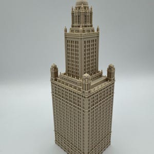 May include: A beige architectural model of a skyscraper with a detailed facade. The building features a tall central tower with a domed top, flanked by smaller towers at the corners. The model is intricately designed with numerous windows and architectural details.