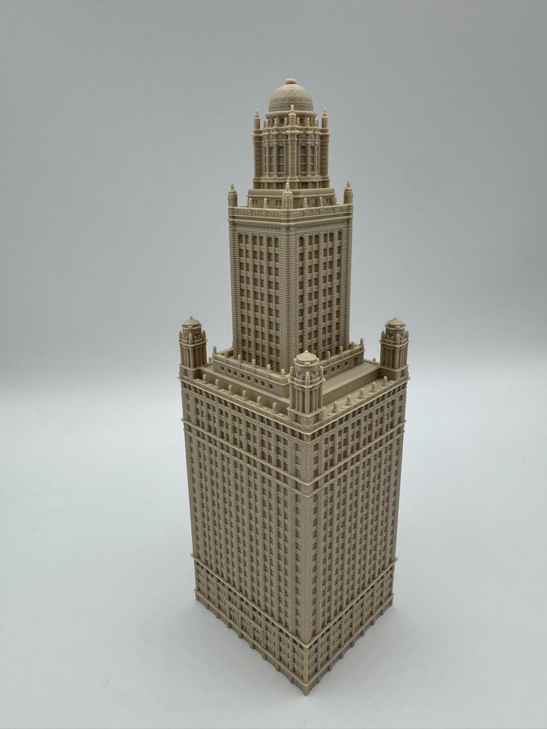 May include: A beige architectural model of a skyscraper with a detailed facade. The building features a tall central tower with a domed top, flanked by smaller towers at the corners. The model is intricately designed with numerous windows and architectural details.
