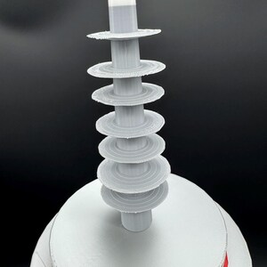 May include: A 3D-printed, abstract sculpture with a white base and a series of gray, circular tiers rising upwards. A central gray column supports the tiers, topped with a red, white, and gray cylindrical element. The sculpture has a modern, geometric design.