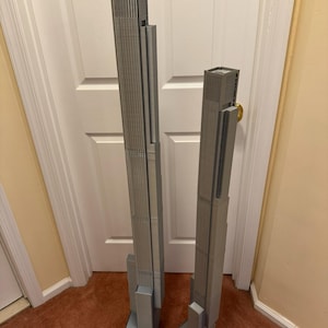 K&ouml;nnte beinhalten: Zwei graue, 3D-gedruckte Modelle von Wolkenkratzern. Das gr&ouml;&szlig;ere Modell ist etwa 61 cm hoch, w&auml;hrend das kleinere etwa 46 cm misst. Beide haben detaillierte Fassaden und rechteckige Sockel, die architektonisches Design zeigen.