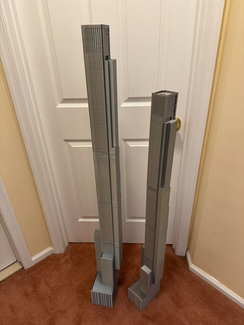 K&ouml;nnte beinhalten: Zwei graue, 3D-gedruckte Modelle von Wolkenkratzern. Das gr&ouml;&szlig;ere Modell ist etwa 61 cm hoch, w&auml;hrend das kleinere etwa 46 cm misst. Beide haben detaillierte Fassaden und rechteckige Sockel, die architektonisches Design zeigen.