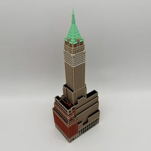 Peut inclure: Une maquette de gratte-ciel composée d'une structure haute, brune et à plusieurs niveaux, surmontée d'un toit pointu vert clair. Le bâtiment présente de nombreux petits détails de fenêtres et une base rectangulaire avec une façade en briques rouge-brun.