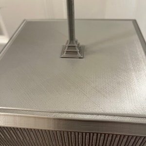 Pu&ograve; includere: Modello grigio stampato in 3D di un monumento. Il modello ha una superficie strutturata e una base dettagliata. La colonna del monumento si eleva da una base quadrata. Il design generale &egrave; geometrico e moderno. Il modello &egrave; probabilmente un oggetto decorativo o uno studio architettonico.