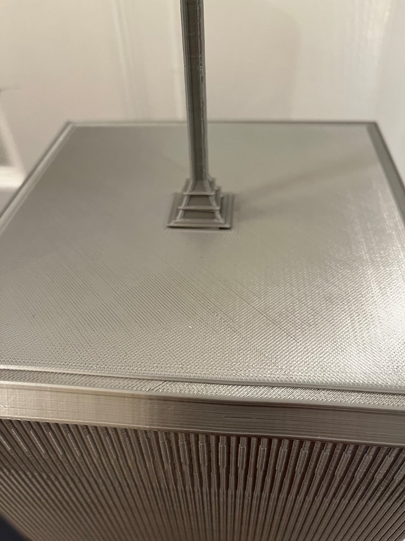 Pu&ograve; includere: Modello grigio stampato in 3D di un monumento. Il modello ha una superficie strutturata e una base dettagliata. La colonna del monumento si eleva da una base quadrata. Il design generale &egrave; geometrico e moderno. Il modello &egrave; probabilmente un oggetto decorativo o uno studio architettonico.