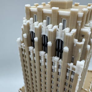May include: A 3D-printed model of a skyscraper, featuring a beige facade with white decorative accents. The model showcases architectural details, including window frames and a textured surface. The building is a miniature representation.