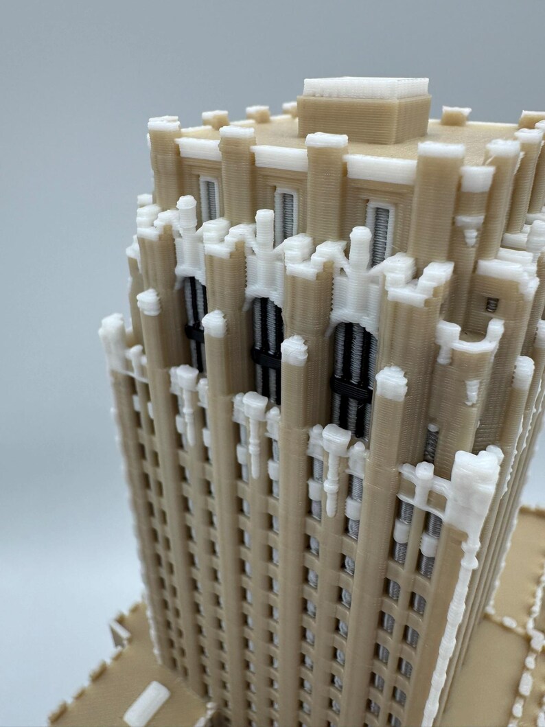 May include: A 3D-printed model of a skyscraper, featuring a beige facade with white decorative accents. The model showcases architectural details, including window frames and a textured surface. The building is a miniature representation.