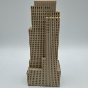 May include: A beige, multi-tiered skyscraper model with a detailed facade of rectangular windows. The building has a stepped design, with the tallest section in the center. The model is likely made of a solid material, and is set against a plain background.