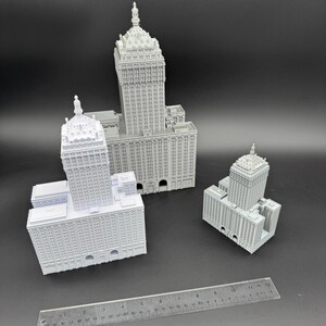 Puede incluir: Tres modelos grises en miniatura de edificios de varios tama&ntilde;os, cada uno con intrincados detalles arquitect&oacute;nicos. Se coloca una regla cerca de los edificios para la escala. Los modelos presentan fachadas detalladas, ventanas y elementos decorativos, mostrando un tema de paisaje urbano.