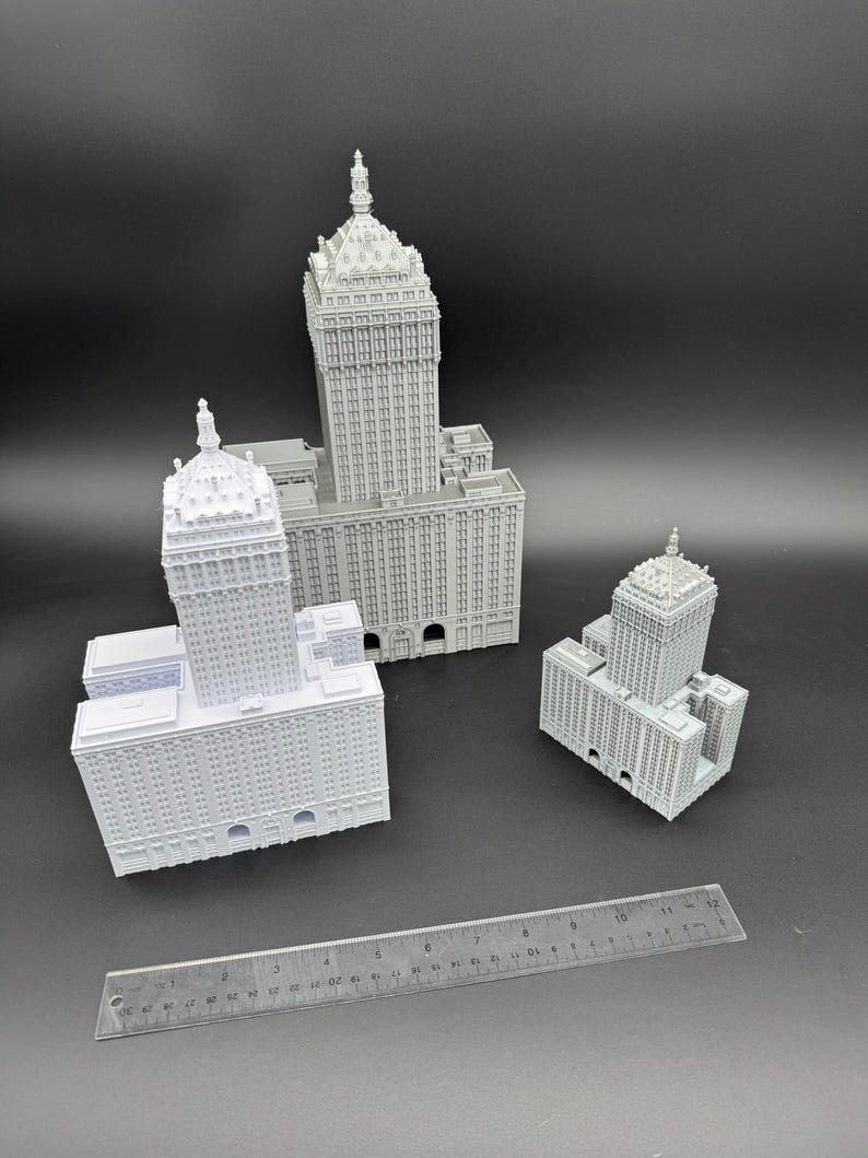 Puede incluir: Tres modelos grises en miniatura de edificios de varios tama&ntilde;os, cada uno con intrincados detalles arquitect&oacute;nicos. Se coloca una regla cerca de los edificios para la escala. Los modelos presentan fachadas detalladas, ventanas y elementos decorativos, mostrando un tema de paisaje urbano.