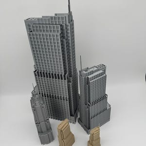 May include: A collection of miniature skyscraper models in various sizes and colors. The tallest buildings are gray, while the smaller ones are tan. A ruler is placed at the bottom, indicating the scale of the models. The buildings have detailed architectural features.