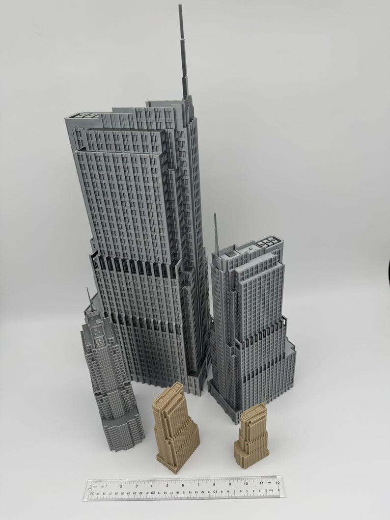May include: A collection of miniature skyscraper models in various sizes and colors. The tallest buildings are gray, while the smaller ones are tan. A ruler is placed at the bottom, indicating the scale of the models. The buildings have detailed architectural features.